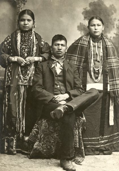 Full-length studio portrait of a Ho-Chunk man sitting and wearing modern dress, flanked by two Ho-Chunk women standing and wearing several necklaces and shawls. They are in front of a painted backdrop. Names (standing, left)) Emma Thunder, Littlesoldier (WePaMaKaRaWinKah) (Daughter of [WaConChaKah] John Thunder aka Dr. Thunder and [WeHonPeKaw] Lucy Bear, Thunder.) (Standing, right) Florence Littlesoldier (MauKonNeeWinKah) (1st wife of Thomas Thunder) (Daughter of [NoGinKah] Little Soldier and Mrs. ChoNeWeKaw.) (Seated) Thomas Thunder (HoonkHaGaKah).