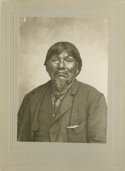 Waist-up studio portrait of an elderly Ho-Chunk man with a beard posing sitting in front of a painted backdrop. He is wearing a suit jacket, vest, and plaid shirt. On the back of the vintage print of this image he is identified as Blackhawk, at 90 years of age.