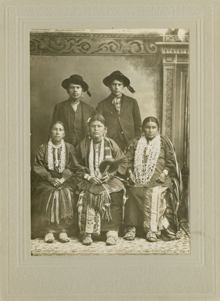 Full-length studio portrait in front of a painted backdrop of three Ho-Chunk women posing sitting in a row, wearing several necklaces, earrings, file bracelets, and shawls wrapped around their waists. Two Ho-Chunk men are posing standing behind them, and wearing suits and hats with drooping eagle feathers. The woman sitting in the center is holding a hand fan.