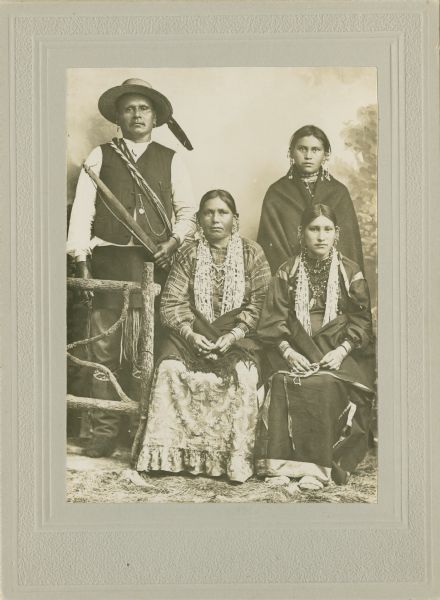 Full-length studio portrait of group posing in front of a painted backdrop with a prop fence. Two Ho-Chunk women are sitting and wearing several necklaces, earrings, and shawl/blankets around their waists. Behind them a Ho-Chunk man, identified as John Thunder, is standing and wearing earrings and a hat with feathers and holding a bow. A young Ho-Chunk woman standing on the right, the wife of Frank Lincoln, is wrapped in a shawl and is wearing several necklaces and earrings. The woman sitting on the left is the wife of John Thunder, and the woman sitting on the right is the wife of Whitegull.