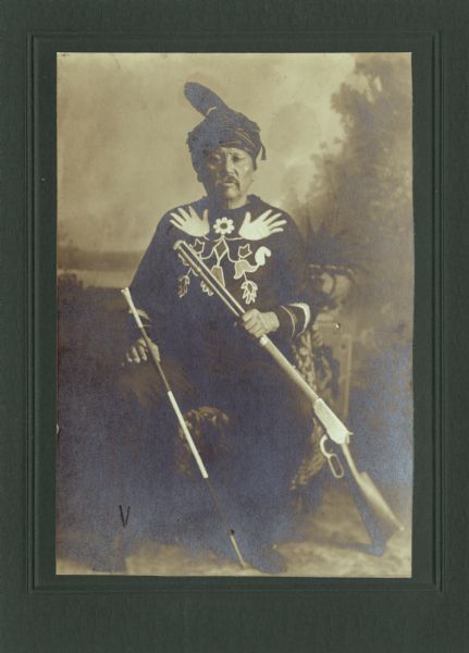 Full-length studio portrait of a Ho-Chunk man, Little Soldier (a.k.a., Strike the Tree, NoGinKah), posing sitting and holding a rifle, and a cane or stick in his left hand in front of a painted backdrop. He is wearing full regalia, including an elaborately tied scarf on his head.