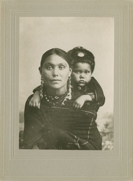 Quarter-length studio portrait of a woman in Ho-Chunk regalia wrapped in a blanket, with a child on her back. The child is wearing a fur hat, and the woman is wearing long earrings and a necklace. They are posing sitting in front of a painted backdrop. Clara Kingsley (Big) Blackhawk (KeesKawWinKah) carries her son Andrew John (Big) Blackhawk (WaConChaHoNoKah) on her back. Using a shawl as a back sling was a common way to carry infants in this period.