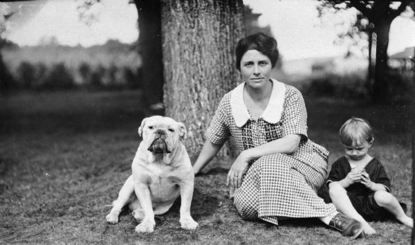 An outdoor portrait of a woman, young child, and a dog, posing sitting on the ground in front of a tree. The woman is sitting in the middle and is wearing a dress, the child is sitting on the right and looking down at something in his hands, and the dog is sitting on the left.