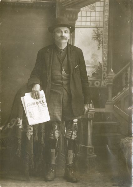 Studio portrait of William Tennant standing and wearing a suit and hat in front of a painted backdrop. He was a newspaper and magazine agent in Black River Falls, Wisconsin, who lost both his legs below the knee. He is facing the viewer and is holding a newspaper in his right hand, and his pant legs have been shortened to reveal his artificial legs.