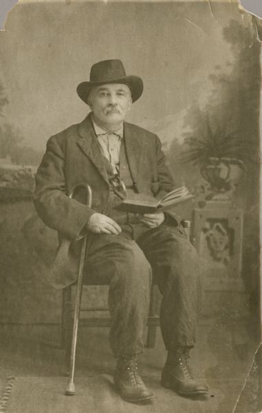 Studio portrait of William Tennant sitting and wearing a suit and hat in front of a painted backdrop. He was a newspaper and magazine agent in Black River Falls, Wisconsin, who lost both his legs below the knee. He is holding an open book in his left hand and a cane under his right arm.