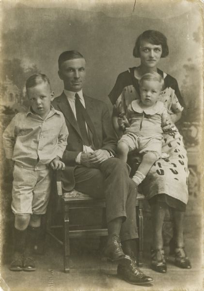 A studio portrait of Shirley Delaney Van Schaick (son of the photographer and Ida Van Schaick), and his family in front of a painted backdrop. Shirley married Rosade or Rosada, and lived in Milwaukee. His two children with Rosada were Edson (b. 1920) and Allen (b. 1924). The woman on the right is Rosada with Allen in her lap (as he was the youngest), Shirley is seated, and young Edson is standing on the left.