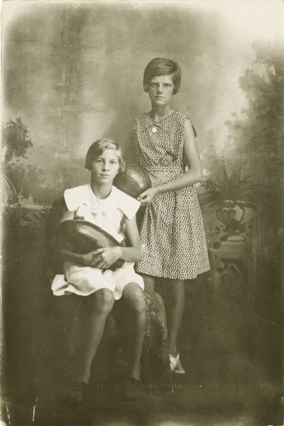 A studio portrait of two young women in front of a painted backdrop. The girl sitting on the left and wearing a white dress is identified as Betty Northup. The girl standing on the right and wearing a patterned dress is identified as Ellen "Dottie" Thompson. Both young women are holding large vegetables in their arms.