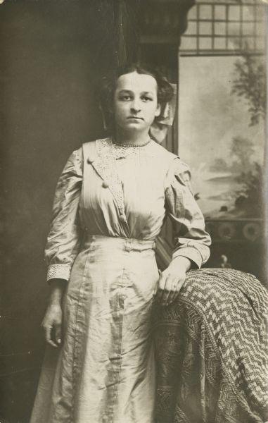 Three-quarter length studio portrait of a young woman wearing a dress standing with her left arm resting on a chair covered with a throw. In the background is a painted backdrop. On the back of the the print, she is identified as "One of the Frieda Schoessers."