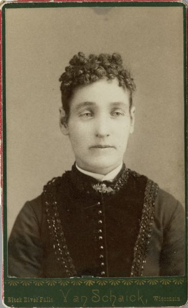 Quarter-length portrait of a woman with dark, curly hair. She is wearing a dress with buttoned bodice with decorative trim, and a collar pin.