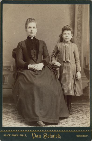Full-length portrait of a seated woman and a standing girl in front of a painted backdrop. The woman is wearing a dark dress with a collar pin, and has her hands in her lap. The girl is wearing a dress and is resting her hand on the arm of the chair.