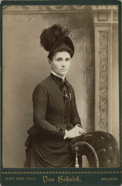 Three-quarter length studio portrait of a woman standing in front of a painted backdrop. She has her hands resting on a velvet upholstered, curved backed chair. She is wearing an intricate dress with many buttons and a clasped neckline, as well as a large hat with feathers.