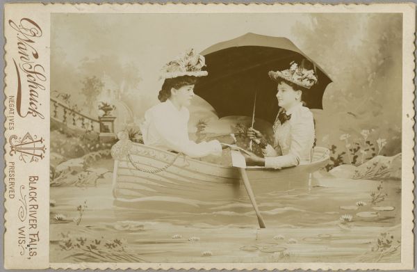 A studio portrait of two women posing sitting in a row boat prop in the middle of painted scenery as the background and foreground. Both women are wearing light-colored and intricate blouses, gloves, and elaborate hats with flowers. The woman sitting on the right is holding an umbrella and a piece of paper. The other woman sitting on the left is holding on to the oars. Neither of the women are looking directly at the camera.