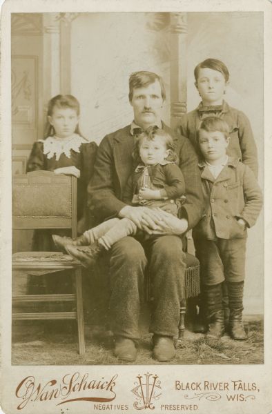 A studio portrait of a man and four children in front of a painted backdrop. The man is sitting on a chair in the center, and is holding one child. Two young boys are standing to the right, and a young girl is standing to the left behind an empty chair.