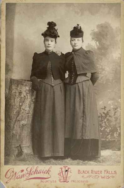Full-length studio portrait of two women standing next to a tall stump in front of a painted backdrop. The woman on the left has one arm resting on the stump and is wearing gloves. Both women are wearing long dresses with buttoned tops, short capes, and elaborate hats.