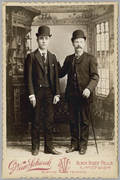 Full-length studio portrait of two men posing standing in front of a painted backdrop. Both men are wearing suits, ties, and hats. The man on the right has a moustache, is holding a cane and resting his right hand on the other man's shoulder.