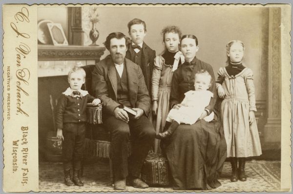 A studio portrait of a family with a man, a woman, and five children, posing in front of a painted backdrop. Both the man and the woman are sitting, and the woman is holding a small child on her lap. Two young girls wearing matching dresses are standing behind the woman. Two young boys are standing near the man, who has a beard and is holding a book in his hands.