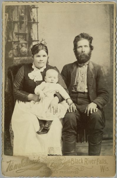 A studio portrait of a man, woman, and infant sitting in front of a painted backdrop. The woman is on the left and is holding the infant in her lap. The man is on the right and has a large beard and moustache. On the back of the print of this image, they are identified as "Pers. & J. family."