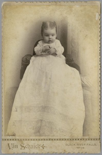 A studio portrait of an infant sitting in a chair, with hands clasped in front of his or her chest, and gazing downward. The infant is wearing a very long white dress with a lace hem, and the hair is styled with a part down the middle. This image is part of the 'Jones Collection,' according to the back of the print.