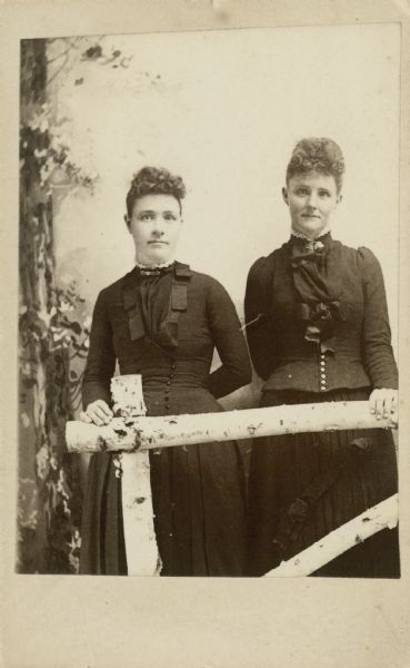 Three-quarter length studio portrait of two women standing in front of a painted backdrop, behind a prop birch fence. Both women are wearing dresses with ruffles and buttons, and have similar hairstyles.