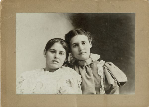 Quarter-length portrait of two young women. Both young women have their hair pulled back and are wearing dresses with lace decoration.