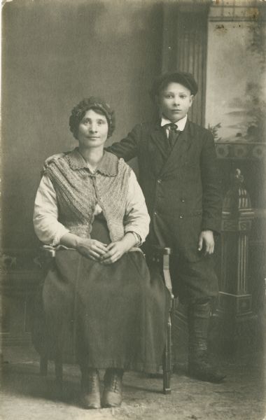 Full-length studio portrait of a woman sitting on the left and a boy standing on the right in front of a painted backdrop. The woman is wearing a knitted hat and vest along with a long skirt and a blouse. The boy is standing with his right arm around the woman's shoulders, and is wearing a suit, tie, and hat.