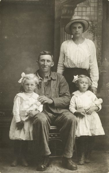 A studio portrait of a young family posing in front of a painted backdrop. A woman is standing on the right in the back, wearing a skirt, ruffled blouse, and hat. In front of the woman is a young girl standing and holding a doll, wearing a dress and a bow in her hair. In the center is a man sitting with his hands resting on the shoulders of another girl standing on the left, who is also holding a doll and wearing a dress and a bow in her hair.