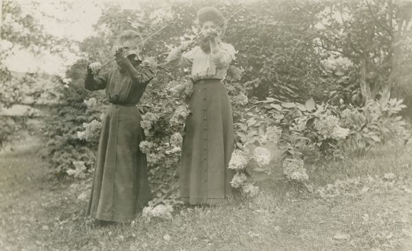 Two women are standing outdoors in front of trees and a blooming bush of flowers, probably hydrangeas, and playing violins. On the back of the print, one of the women is identified as Mollie Ma??llhe.