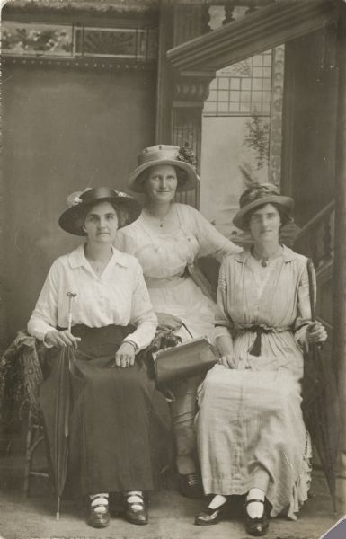 Full-length studio portrait of three women sitting in front of a painted backdrop. Each woman is wearing an elaborate hat and a necklace. The woman sitting on the left is holding an umbrella, the woman in the center is holding a purse, and the woman on the right is wearing eyeglasses and is holding an umbrella.