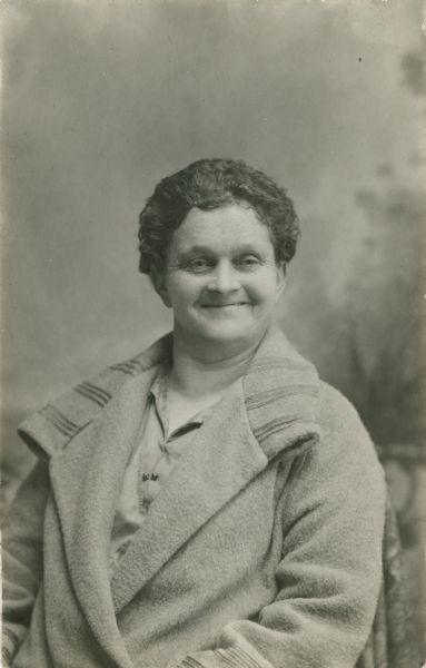 Waist-up studio portrait of a smiling woman sitting in front of a painted backdrop. She is wearing a coat and has short, curly hair.