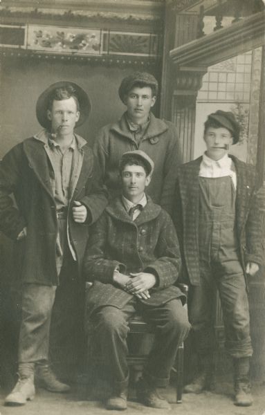 Full-length studio portrait of four young men in front of a painted backdrop. One young man is sitting in the center, with the other three young men standing around him. Each man is wearing a coat and a hat, and two of them have pipes in their mouths.