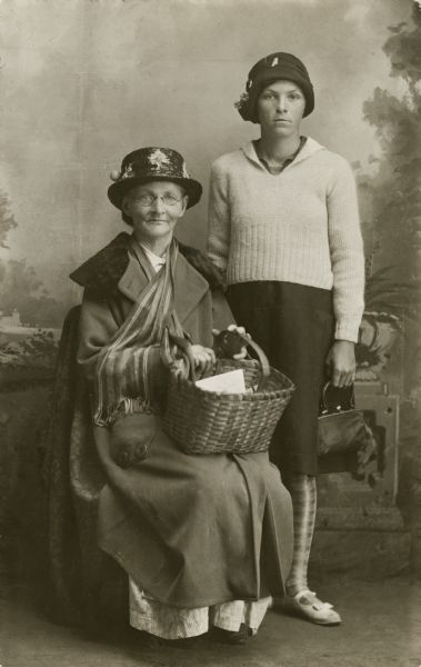 Full-length studio portrait of two women posing sitting and standing in front of a painted backdrop. On the left, the older woman is holding a basket on her lap, and is wearing a long coat, a scarf, glasses, and an embellished straw hat. The younger woman is standing on the right, wearing a sweater, skirt, patterned tights, a hat, and is carrying a purse.