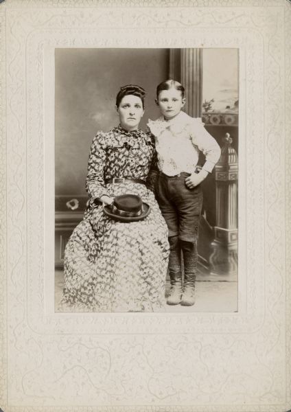 A studio portrait of a woman and a young boy posing in front of a painted backdrop. The woman is sitting on the left wearing a patterned dress and holding a hat on her lap. The young boy is standing on the right and is wearing a light-colored shirt with a ruffled collar.