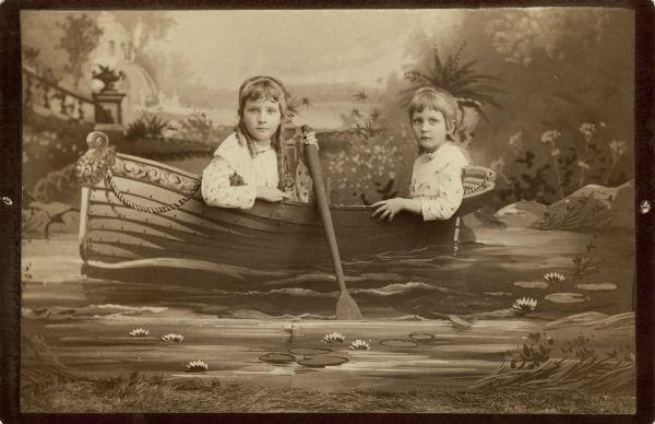 A studio portrait of two young girls posing sitting in a painted rowboat prop in front of a painted backdrop. Both girls are wearing printed dresses and are looking directly at the camera.