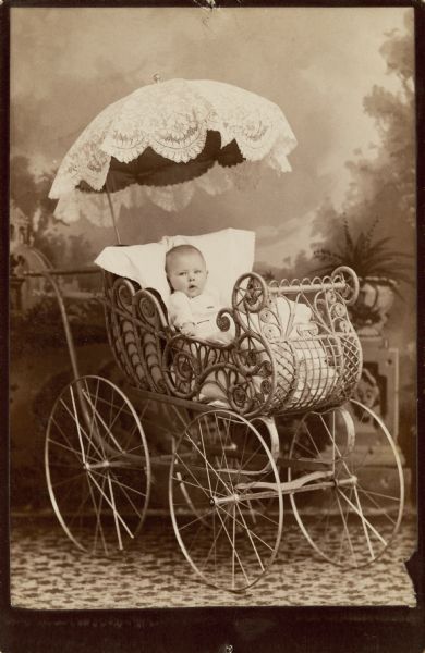 A studio portrait of an infant in an elaborate stroller/carriage/buggy in front of a painted backdrop. The stroller is covered by a lacy umbrella, and the basket part of the stroller seems to be made out of intricately curved wicker, and is filled with blankets and a pillow for the infant to rest on. In addition, the stroller has four large metal wheels.