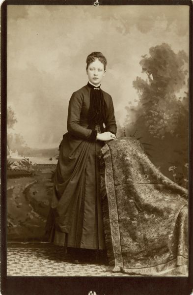 Full-length studio portrait of a woman standing in front of a painted backdrop. She is wearing a long dress with velvet accents, a bracelet, a ring, and a collar pin. She has her hands placed on a cloth-covered chair on the right.