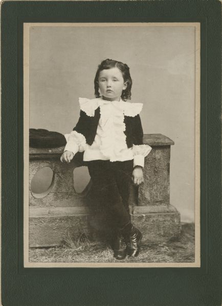 Full-length studio portrait of a young child leaning against a prop stone wall. The child is wearing dark pants, and a white and ruffled shirt with dark accents, and has dark, chin-length, curly hair. A matching hat is atop the stone wall next to the child, who has a nonchalant expression on his or her face.