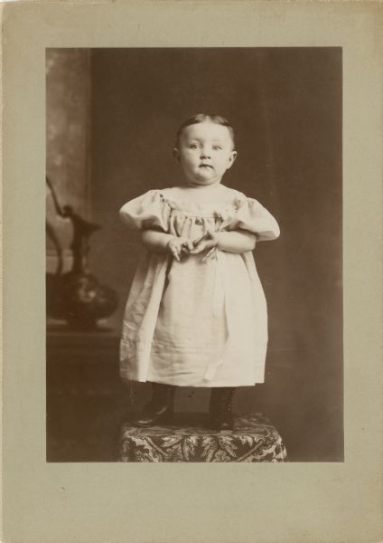 A studio portrait of a baby standing on a cloth-covered surface in front of a painted backdrop. The baby is wearing a long, light-colored dress and boots.