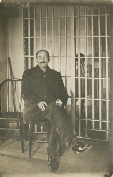 Copy photograph of a man posing sitting in jail, probably John Deitz in Hayward. He appears to have a cast or bandage on his left hand. A young girl is standing in the background behind the bars.