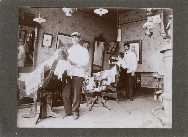 The interior of a barbershop with customers in chairs and barbers at work. Numerous mirrors, pictures, and lamps are hanging from the walls and ceiling.