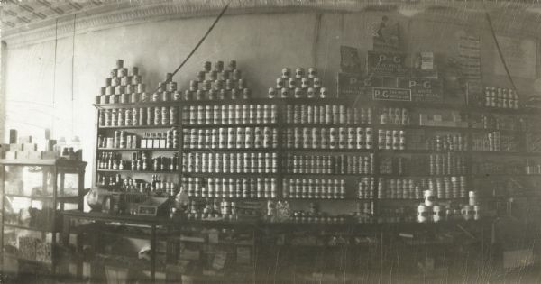 The interior of a store, with a glass counter and shelves full of stacked canned goods. On top of the shelves on the right is a display advertising: "P and G The White Naphtha Soap."