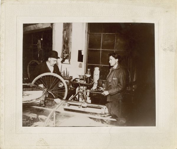 Two men in a repair shop, probably the Repair Shop of Leslie Werner. Leslie Werner is identified as the man wearing the hat on the right.
