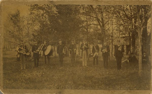 Group portrait of nine men standing and two men sitting in a field with trees in the background. Each standing man is holding a musical instrument, including drums, a tuba, and trumpets. The other two men are sitting against trees on the right.