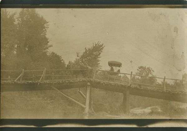 View from below of two horses pulling a carriage with one person in it  traveling over a wooden bridge.