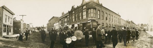 A panoramic view of a Memorial Day parade/celebration as it rounds the corner of a street. According to text on the back of the print, this celebration took place after 1906, and down the street on the left side is a 'homecoming arch.' The text also identifies some of the participants as "Irving Hoften Hagen snare drums, C.W. Reiels Base Drum, ?auera Brevey in front of drums with cap, John Parnell Civil War Vet behind drums."