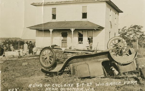 A view of the ruins of a cyclone, showing a roofless house and an overturned automobile in the yard. Several people are standing in the yard, and two children are standing on the porch of the house near two broken windows. Text on front reads: "Wm. Brace Farm near Bloomer, Wis.," and typewritten text on back reads: "This "Lizzie" will be allright with a new engine and three new wheels. Less than $1.00 would have carried a windstorm policy."