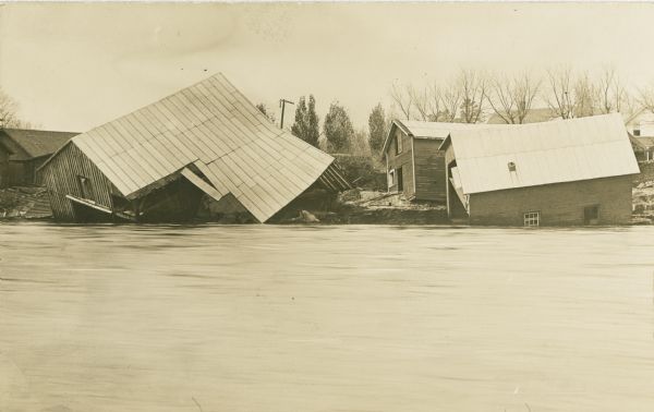 View across river of several collapsing buildings on the edge of flooded river banks, during the flood.