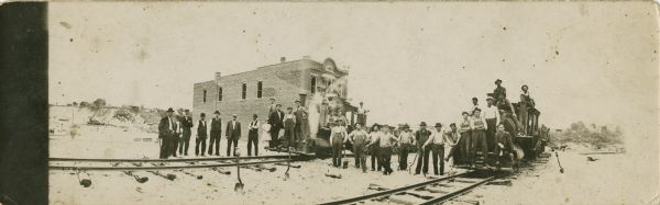 Panoramic view of group of men, boys, and girls lined up along railroad tracks, posing sitting and standing on and near two trains. There is a brick building in the background.