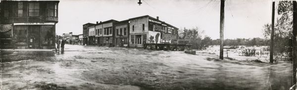 A panoramic view of Black River Falls flooded streets, possibly taken during the flood of 1911. Three men are standing on the sidewalk next to the "Tollack & Son" storefront on the left street corner.