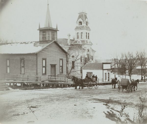 View from across street of a dentist office and Baptist Church along a road. Two horse-drawn carriages are being driven in opposite directions on the road.