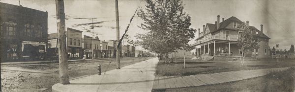 Panoramic view of a street corner. Commercial buildings are along the street on the left, including D.W. Corning Groceries. A large group of people are on the wrap-around porch of the Hotel Washburn, which is on the corner lot on the right. A group of men are standing on the sidewalk.